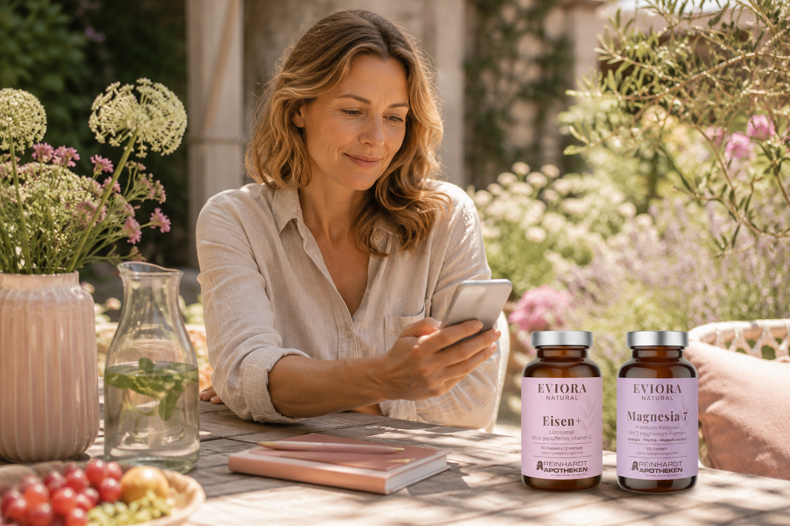 Frau sitzt an einem sommerlichen Gartentisch und blickt auf ihr Smartphone. Daneben stehen die EVIORA Produkte Eisen+ und Magnesia 7 in heller, natürlicher Umgebung.